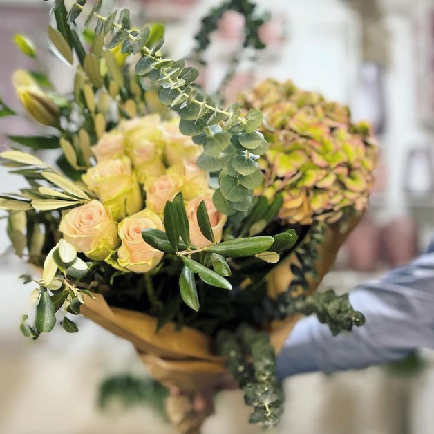 En hånd holder en stor blomsterbuket bestående af lyserøde roser, grønne planter og eukalyptus. Buketten er indpakket i brun papir og præsenteret i et lyst miljø.