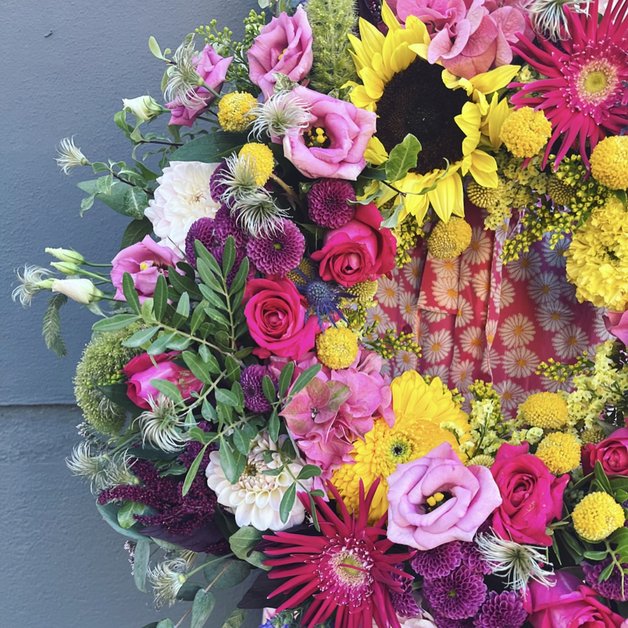 En farverig blomsterkrans sammensat af roser, solsikker, georginer og forskellige grønt. Blomsterne har nuancer af pink, gul, hvid og lilla, omgivet af en lys baggrund med et blomstermønster. Kransen giver et livligt og festligt indtryk.