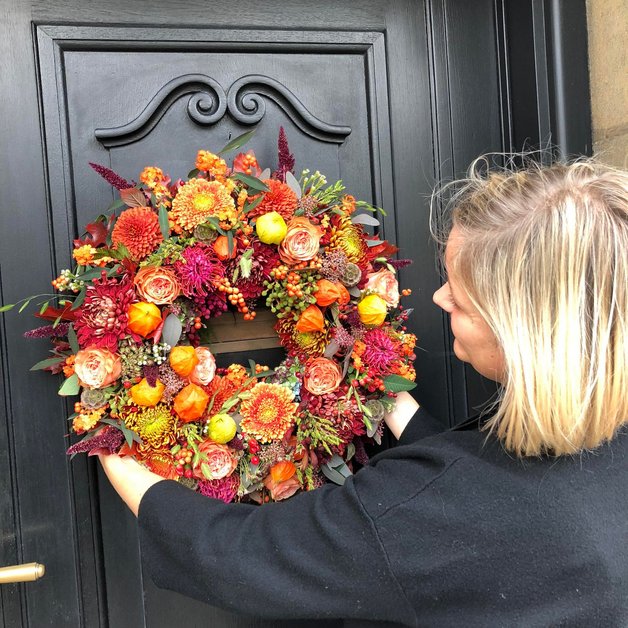 En person hænger en farverig krans lavet af blomster og frugter op på en dør. Kransen indeholder røde, orange og grønne nuancer, og personen fokuserer på at placere den korrekt. Døren er sort og har en enkel stil.
