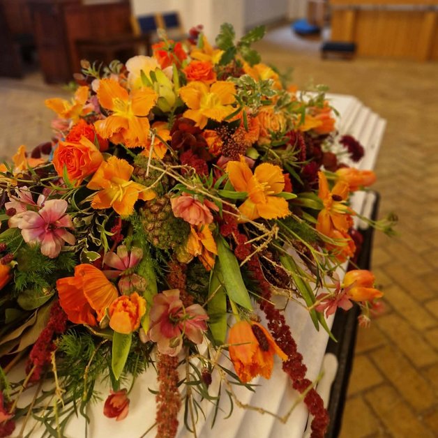 Kistedekoration orange/ efterår En kistedekoration i varme orange og røde nuancer pryder en hvid kiste i en kirke. Baggrunden viser en simpel kirkelig indretning med en alterplads og bænke.