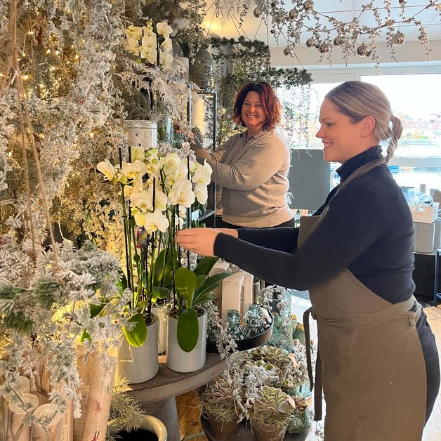 Juleoppyntning To kvinder står i en blomsterbutik, omringet af frostede dekorationer. Den ene kvinde placerer en orkidé i en potte, mens den anden arbejder med en blomsterarrangering. Butikken har en hyggelig og vinterlig atmosfære.